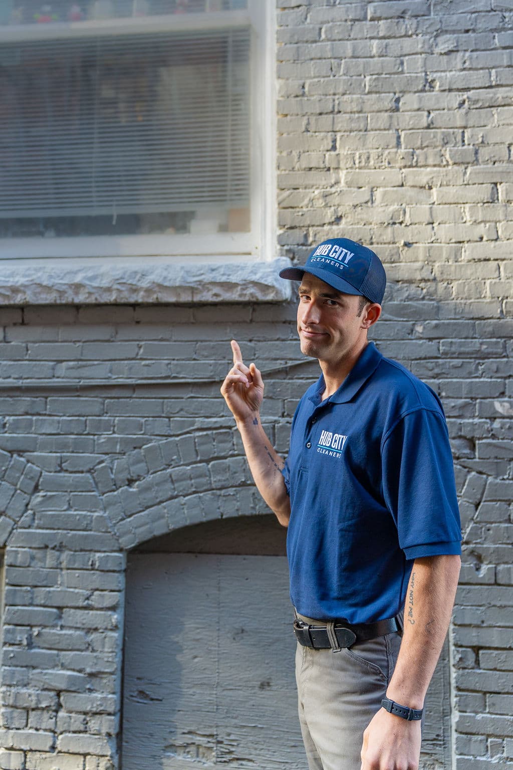 Caleb in a Hub City Cleaners polo and cap, pointing up at a storefront window